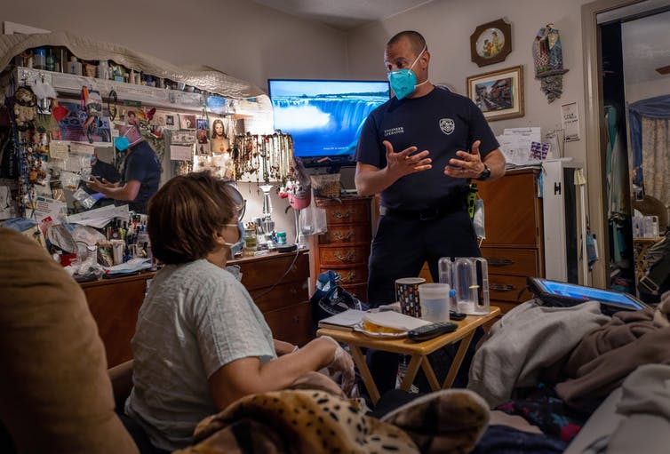 A man in a blue uniform who is wearing a mask speaks to a woman sitting in a recliner in a living room.