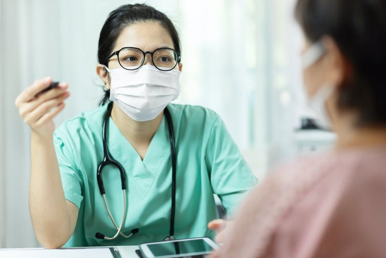 Masked health care provider in green scrubs talking to patient.