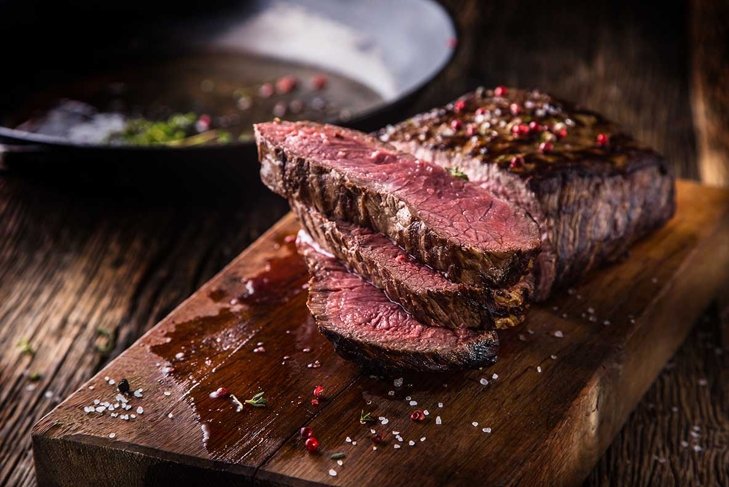 Juicy medium Beef Rib Eye steak slices in pan on wooden board with fork and knife herbs spices and salt.